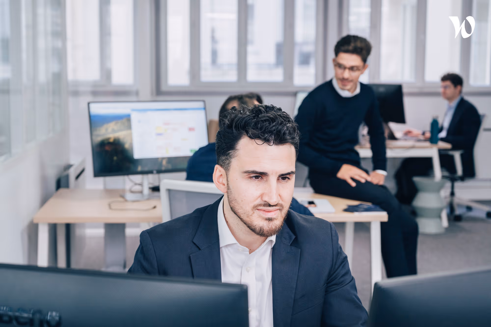 Homme en costume regardant deux écrans d’ordinateur dans un bureau moderne avec collègues en arrière-plan.