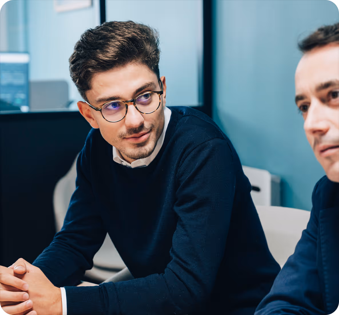 Deux hommes en réunion de travail, l'un portant des lunettes et un pull noir, l'autre en costume bleu.