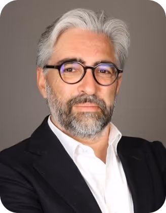 Homme aux cheveux gris et barbe, portant des lunettes rondes, veste noire et chemise blanche.