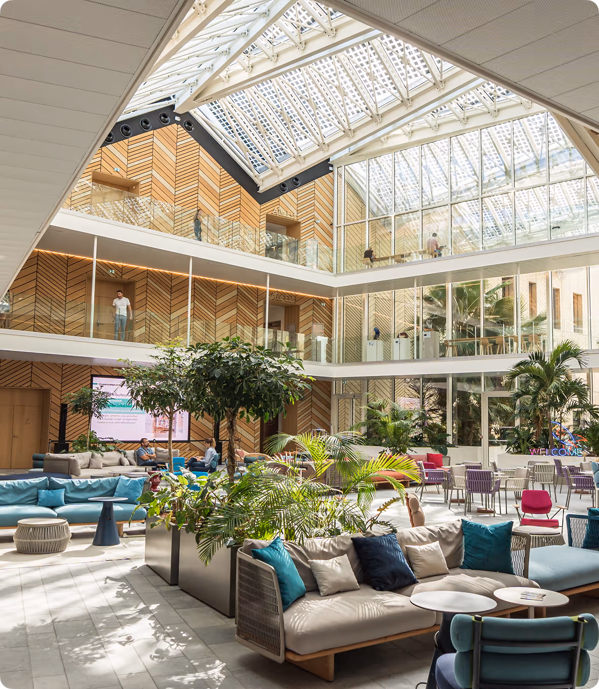 Espace commun ouvert avec canapés, plantes, tables et murs en bois sous un toit en verre.