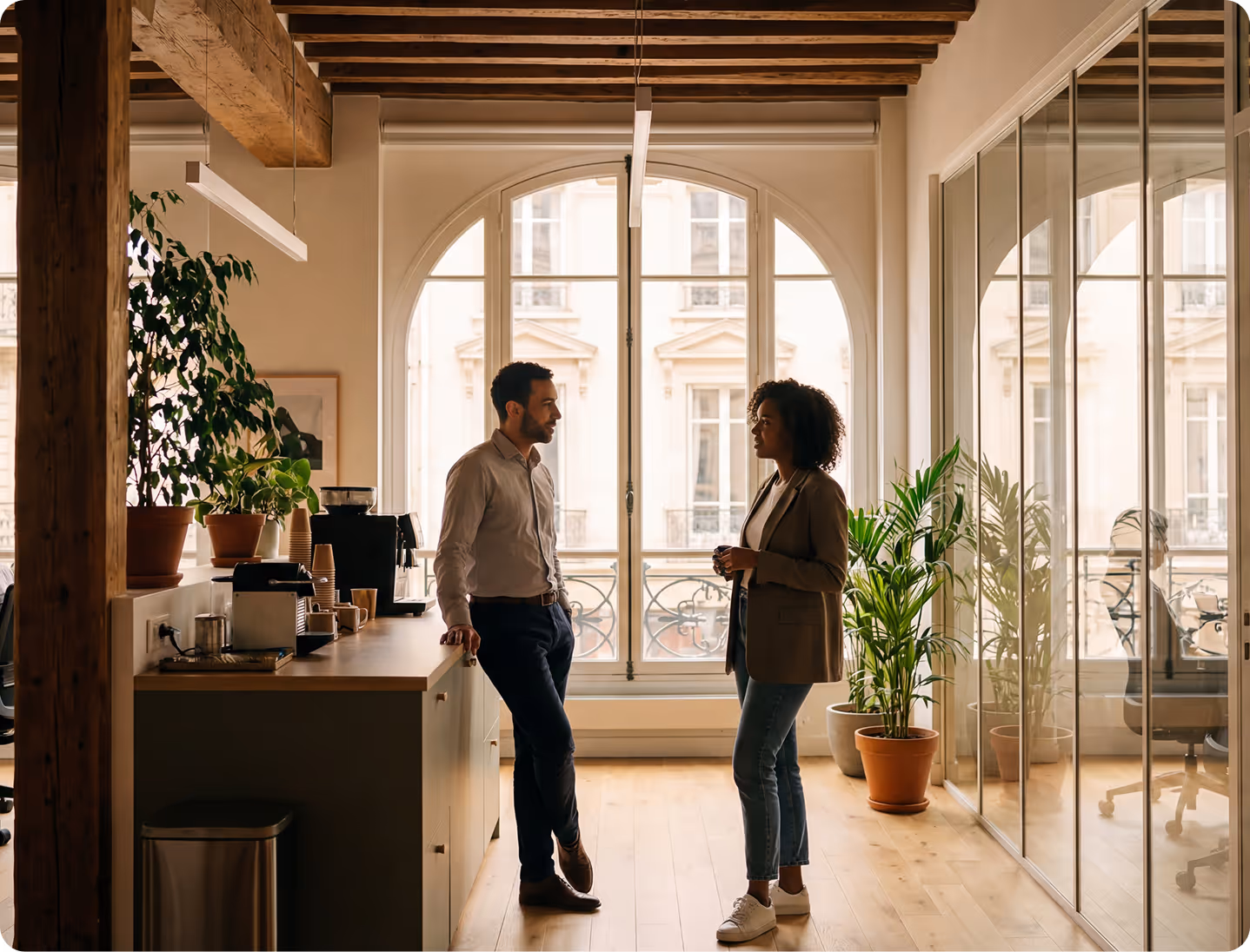 Deux collègues discutent dans un bureau lumineux avec plantes et grandes fenêtres cintrées.