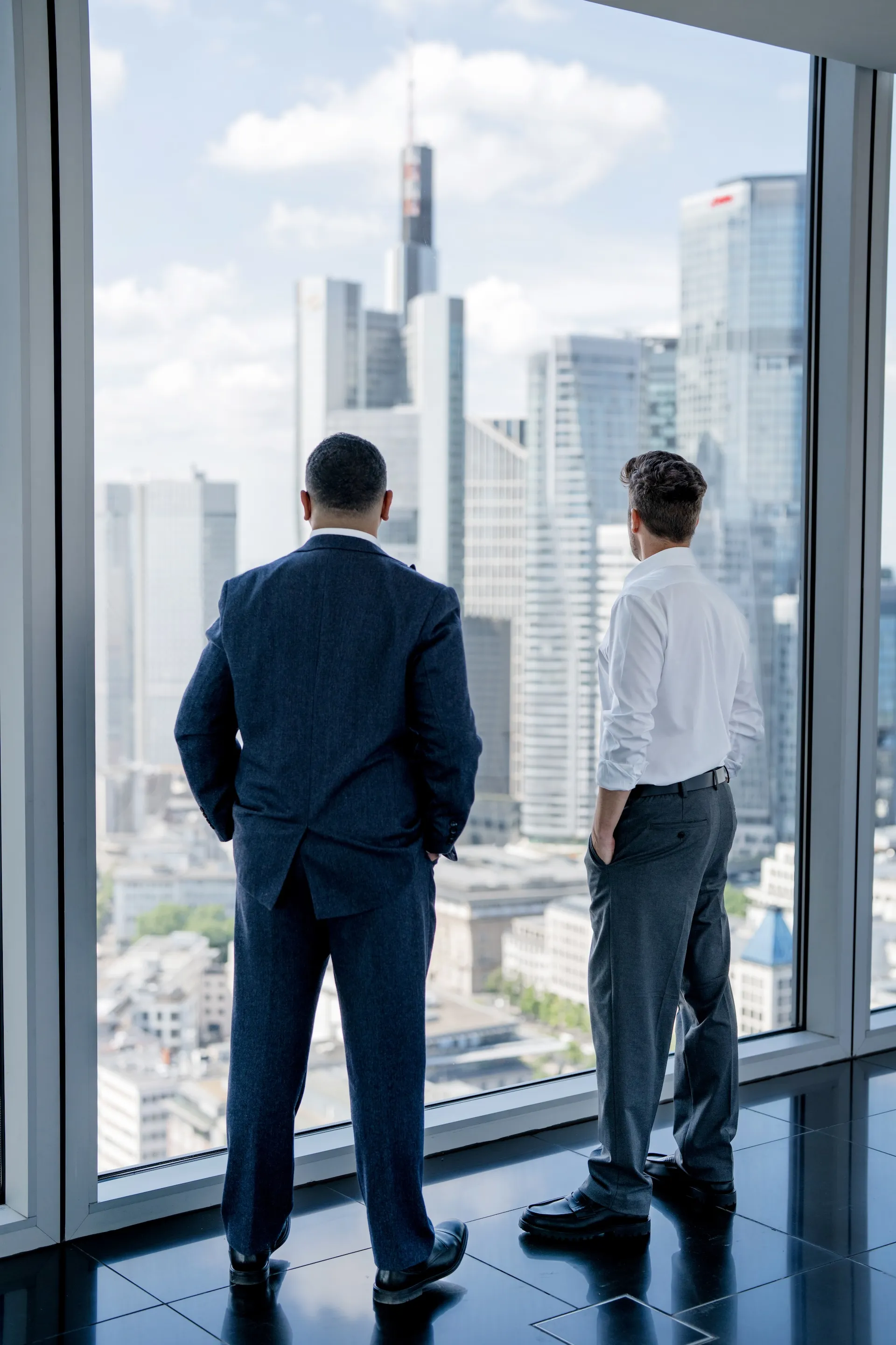 Fabio Waldrón Pelegrin und Sascha Reese im Anzug stehen vor einem großen Fenster mit Blick auf eine moderne Stadtlandschaft.