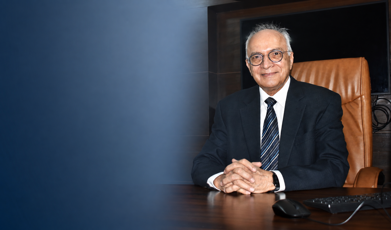 Smiling elderly man in a suit and tie seated at a desk with a keyboard and mouse.