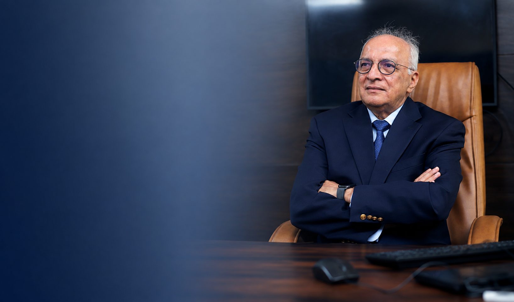 Older man in glasses and navy suit sitting with arms crossed at a desk in an office.