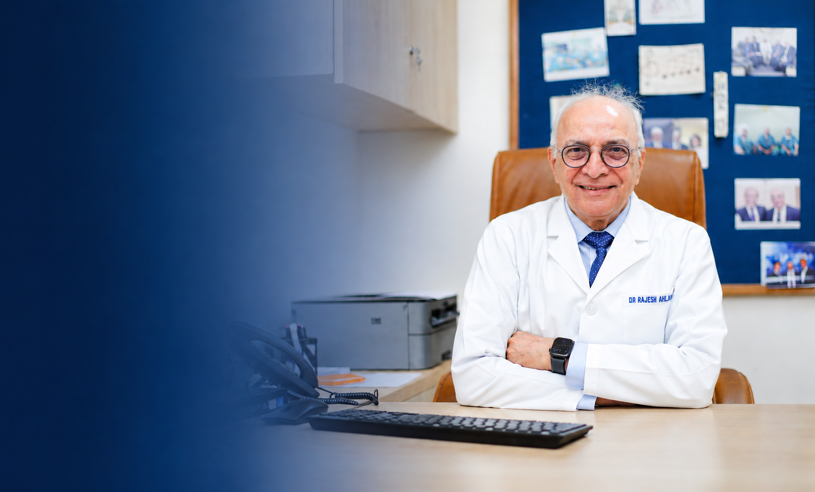 Smiling male doctor with glasses and white coat sitting at a desk in an office with photos on wall.