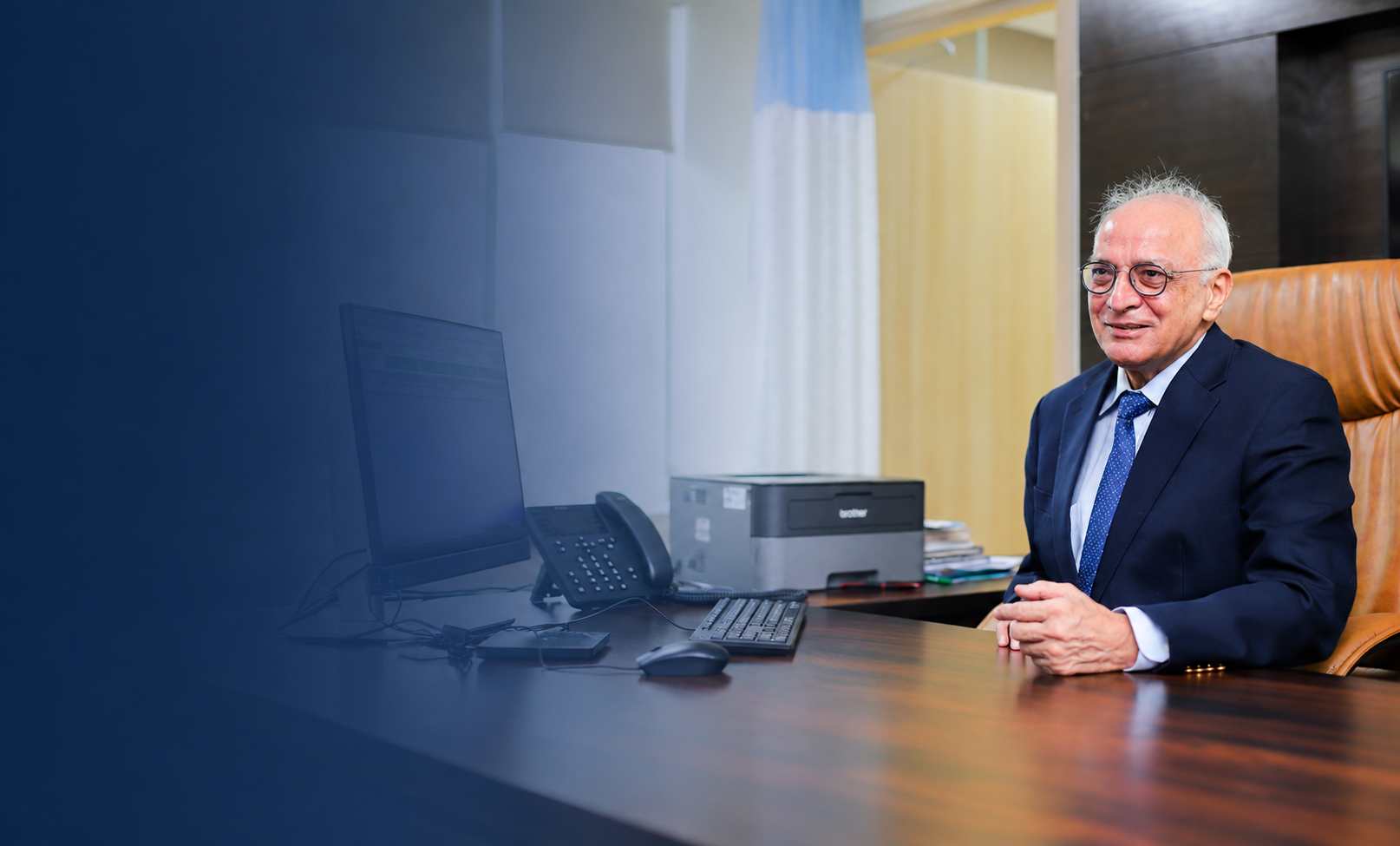 Elderly man in a suit and glasses smiling while sitting at an office desk with computer and printer.