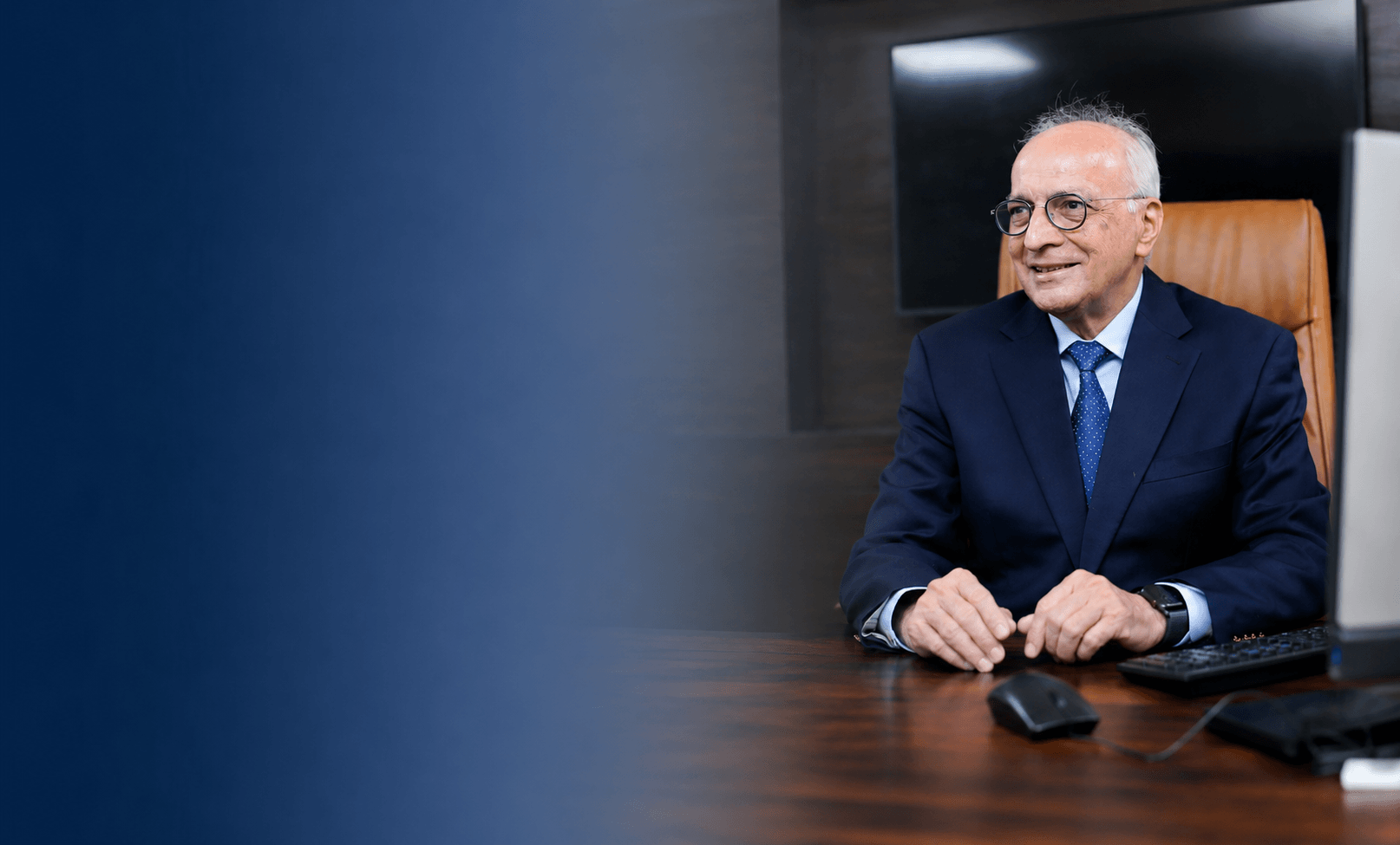 Elderly man in a navy suit and glasses sitting at a desk with a computer mouse and keyboard.