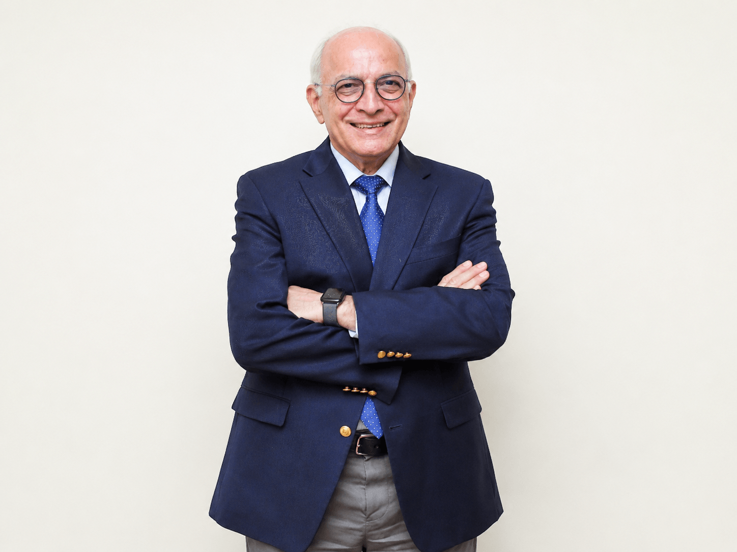 Smiling older man in glasses, navy blazer, blue polka dot tie, and smartwatch stands with arms crossed.