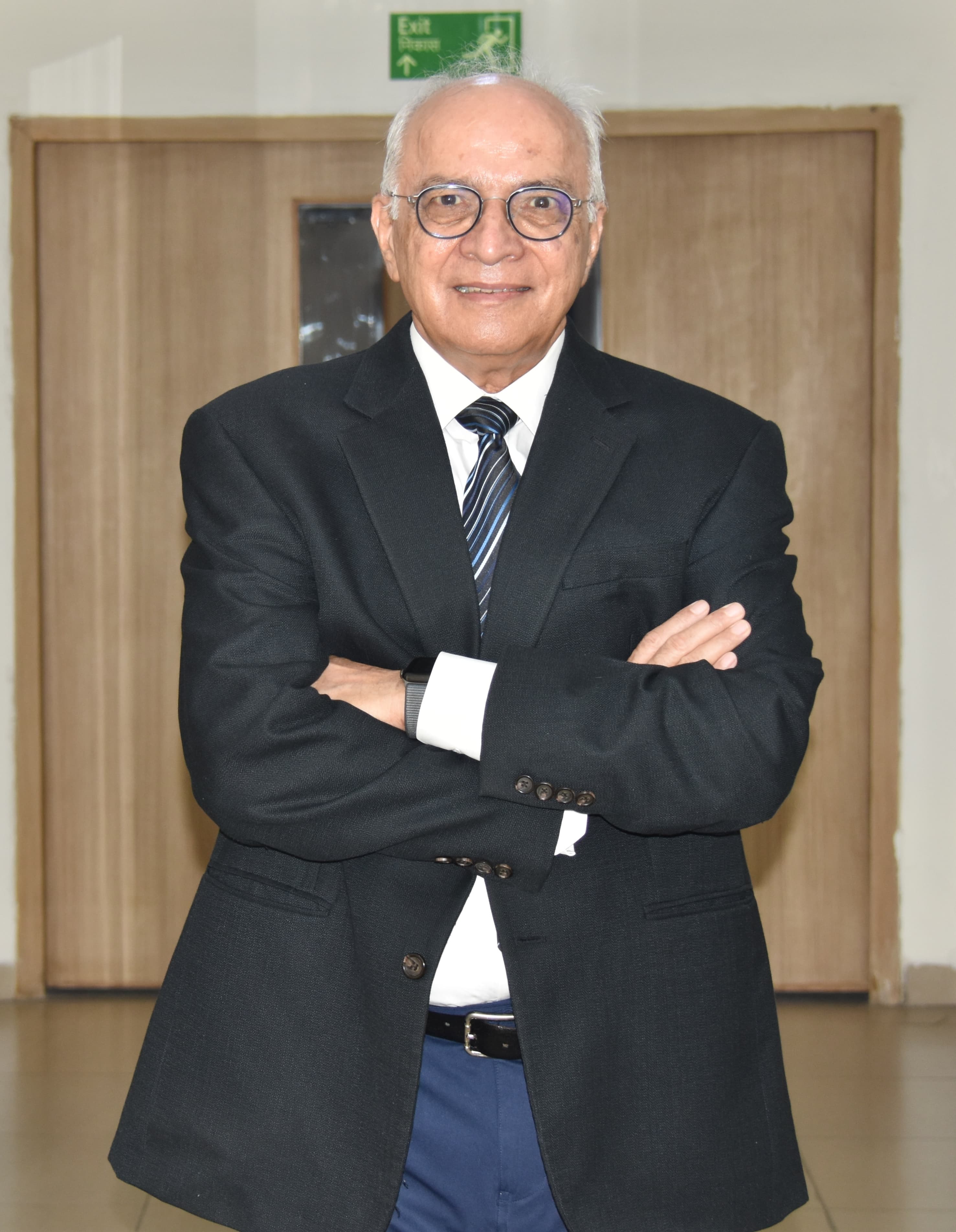 Smiling elderly man with glasses in black suit crossing arms indoors in front of wooden doors.