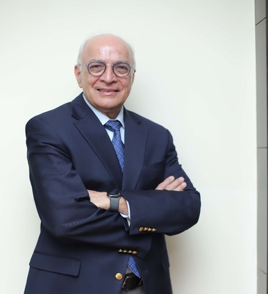 Smiling older man with glasses in a dark suit and blue tie standing with arms crossed against a wall.