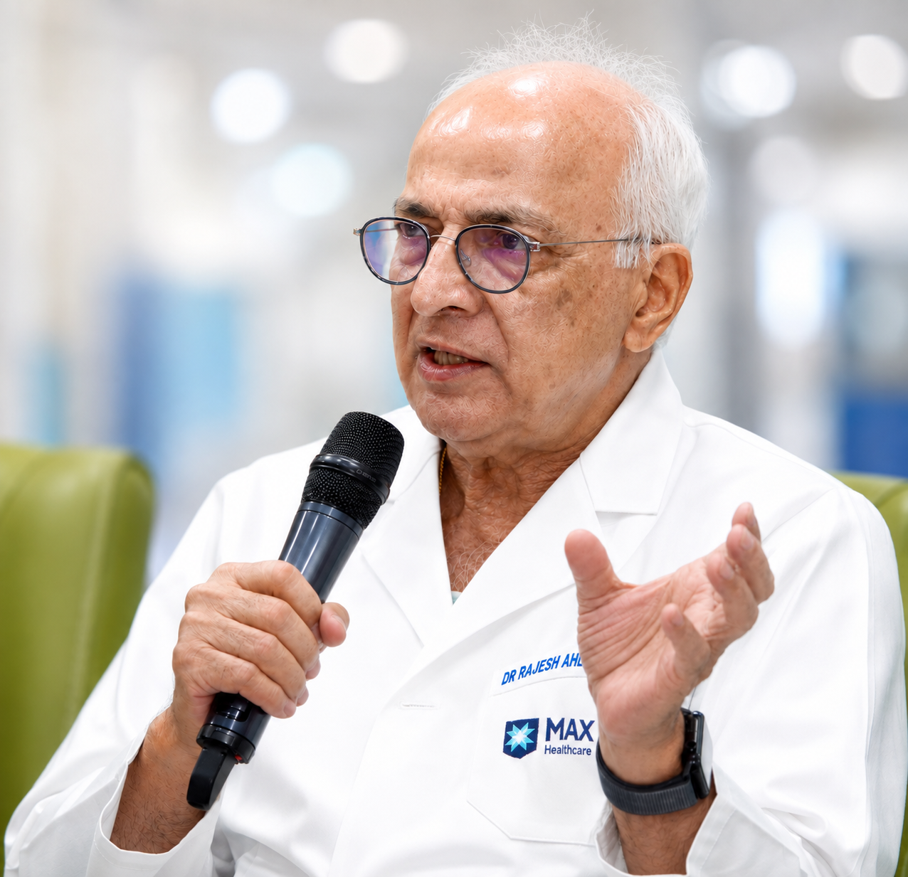 Elderly male doctor in white coat speaking into microphone with expressive hand gesture.