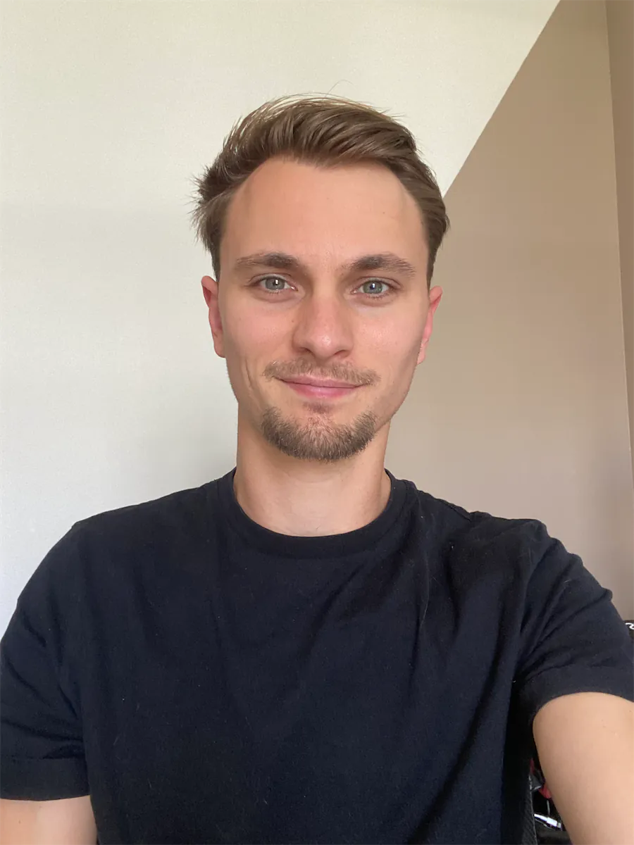 Homme souriant portant un t-shirt noir devant un mur blanc et beige.
