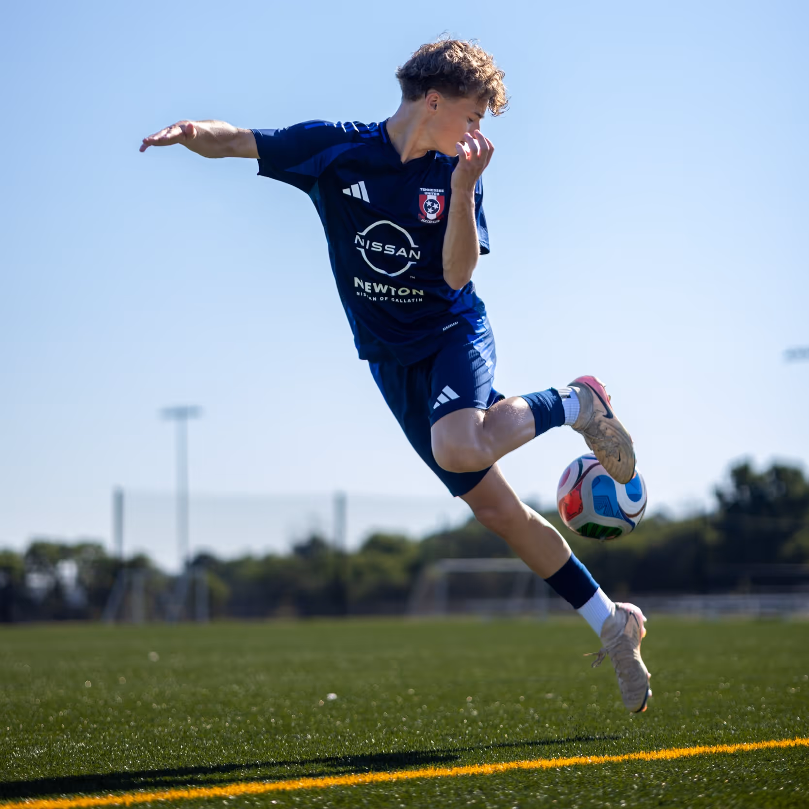 Weston Ochs in blue uniform mid-air kicking a ball on green field near yellow line.
