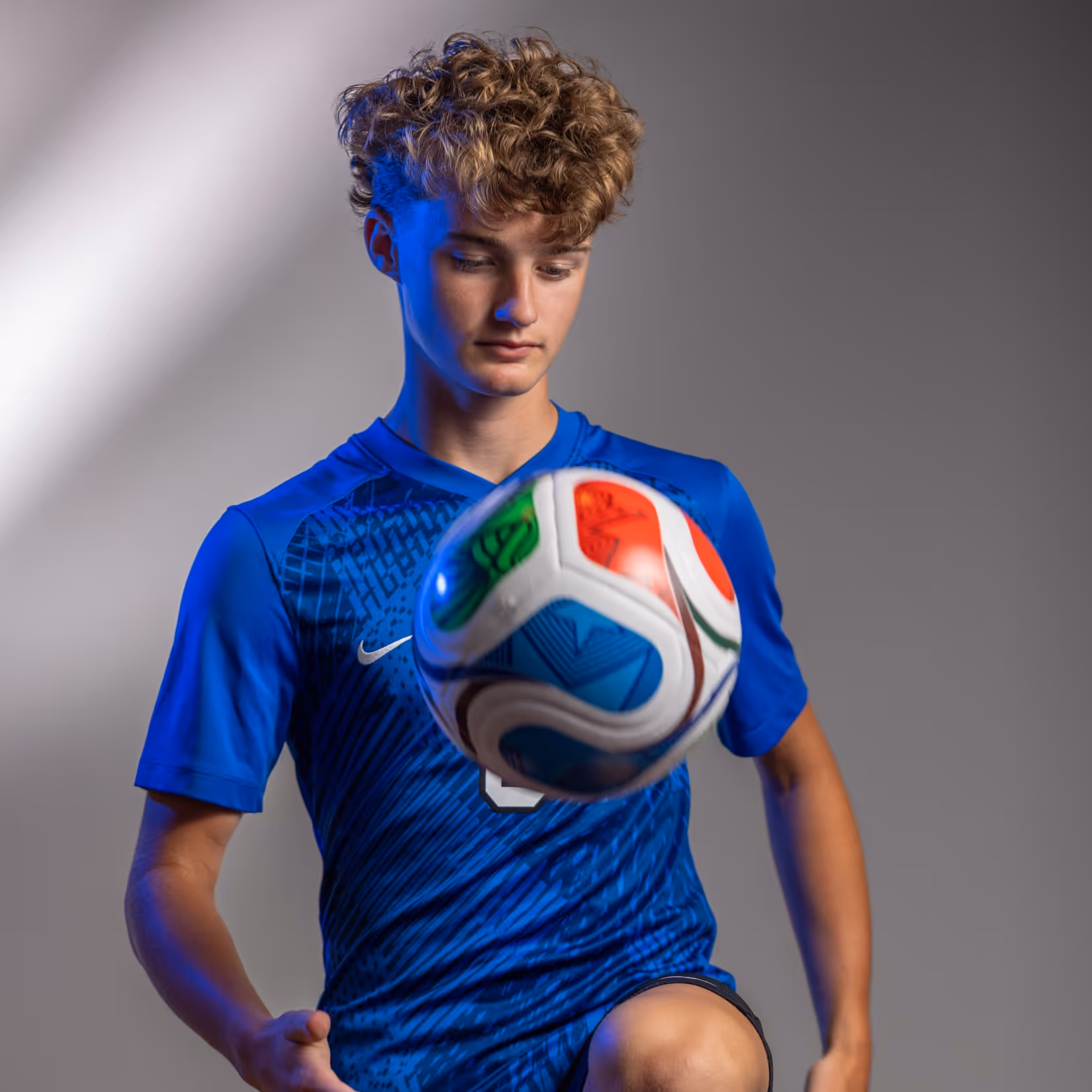 Weston Ochs in blue sportswear balancing a colorful soccer ball on his knee against gray background.