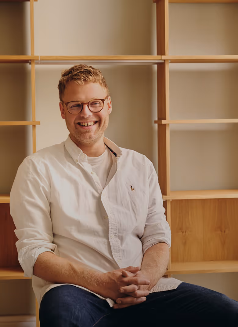 Smiling man with glasses wearing a white shirt, seated in front of empty wooden shelves.
