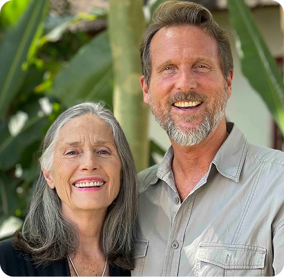Smiling middle-aged woman with long gray hair standing next to smiling middle-aged man with short hair and beard, both in casual clothing, with green foliage in the background.
