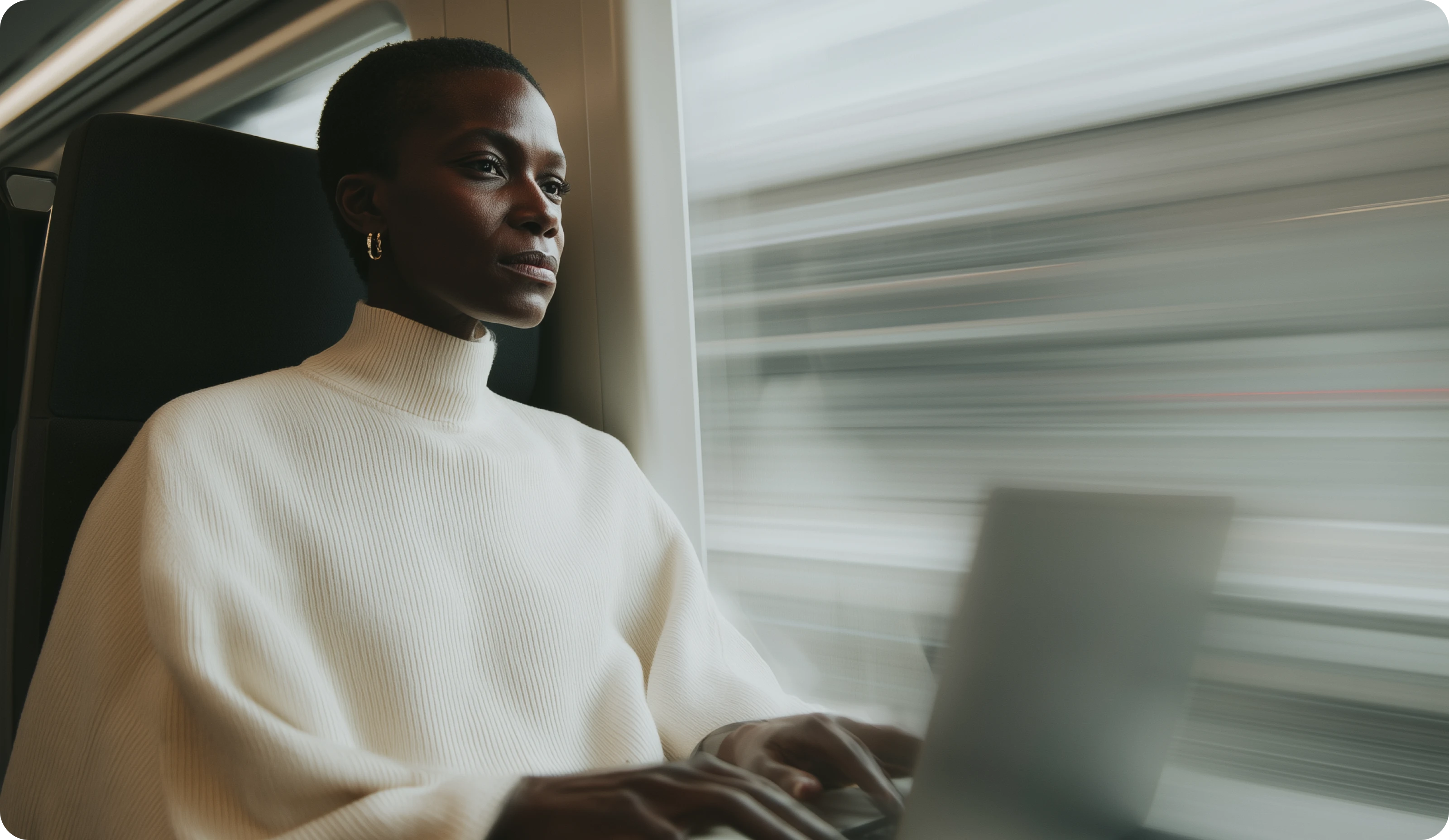 Woman in white turtleneck using laptop while gazing out a fast-moving train window.