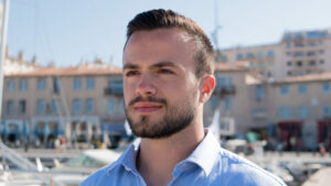 Jeune homme barbu portant une chemise bleue, en extérieur avec un port de plaisance et des bâtiments en arrière-plan.