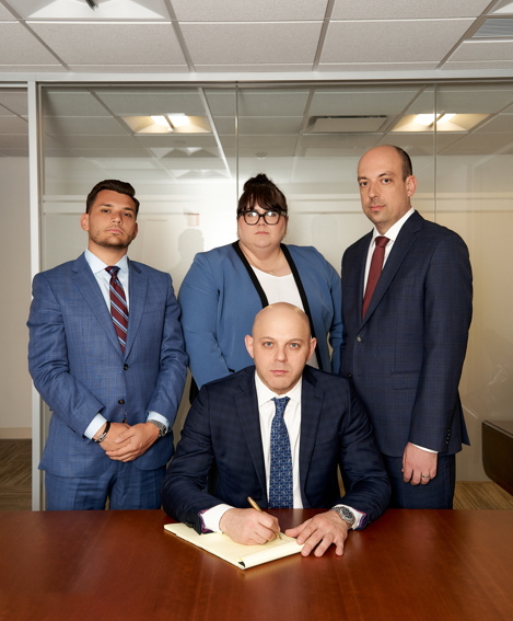Four business professionals in suits posing in an office, one seated at a desk writing in a notebook.