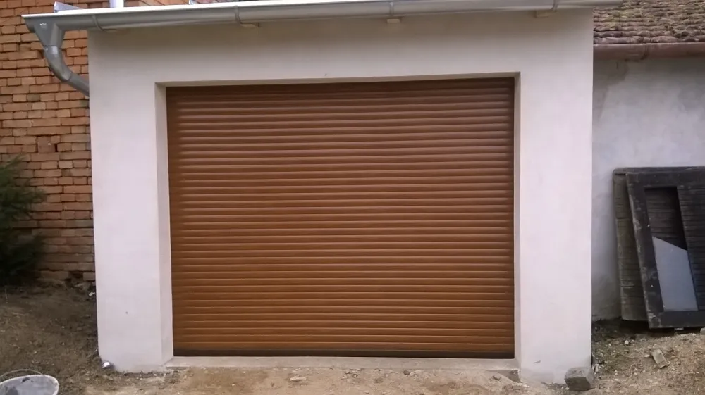 Hnědá rolovací garážová vrata v bílé zdi venku u domu s cihlovou zdí vlevo.