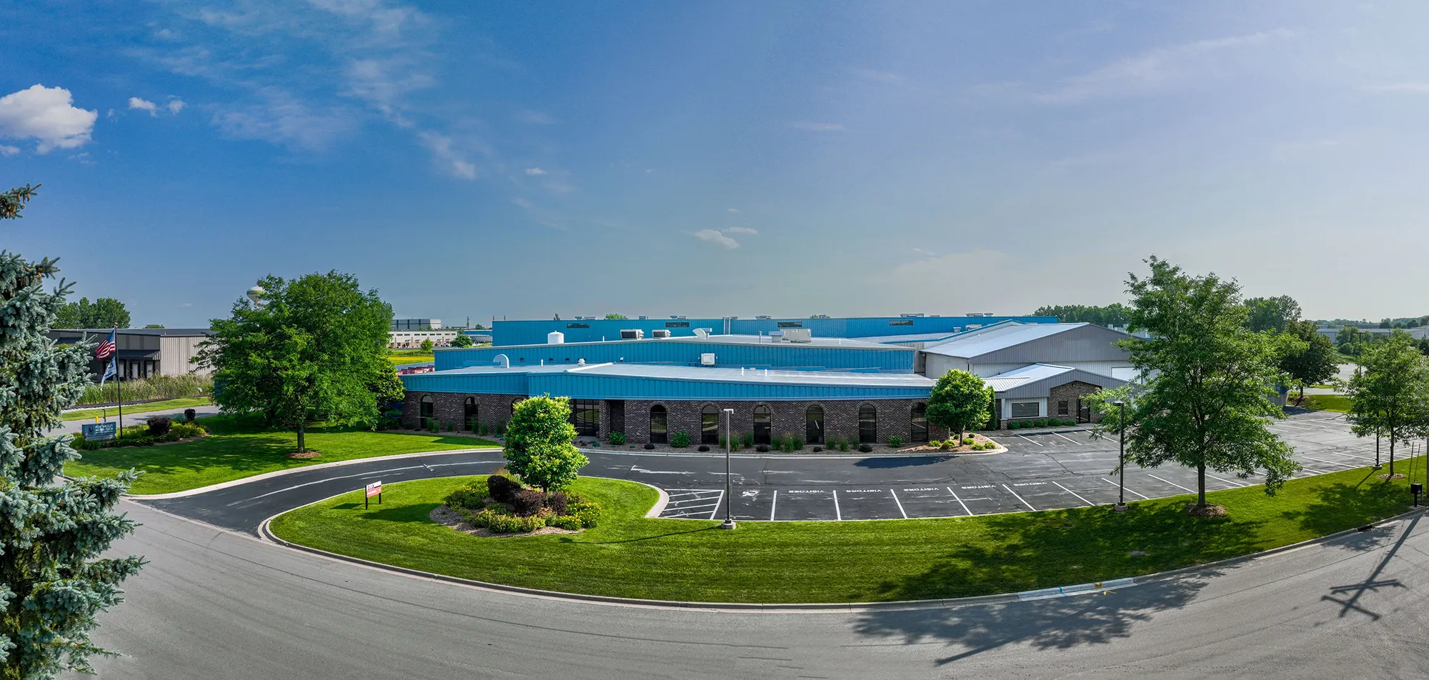 A large industrial building with blue roofs, surrounded by green trees and empty parking lots.