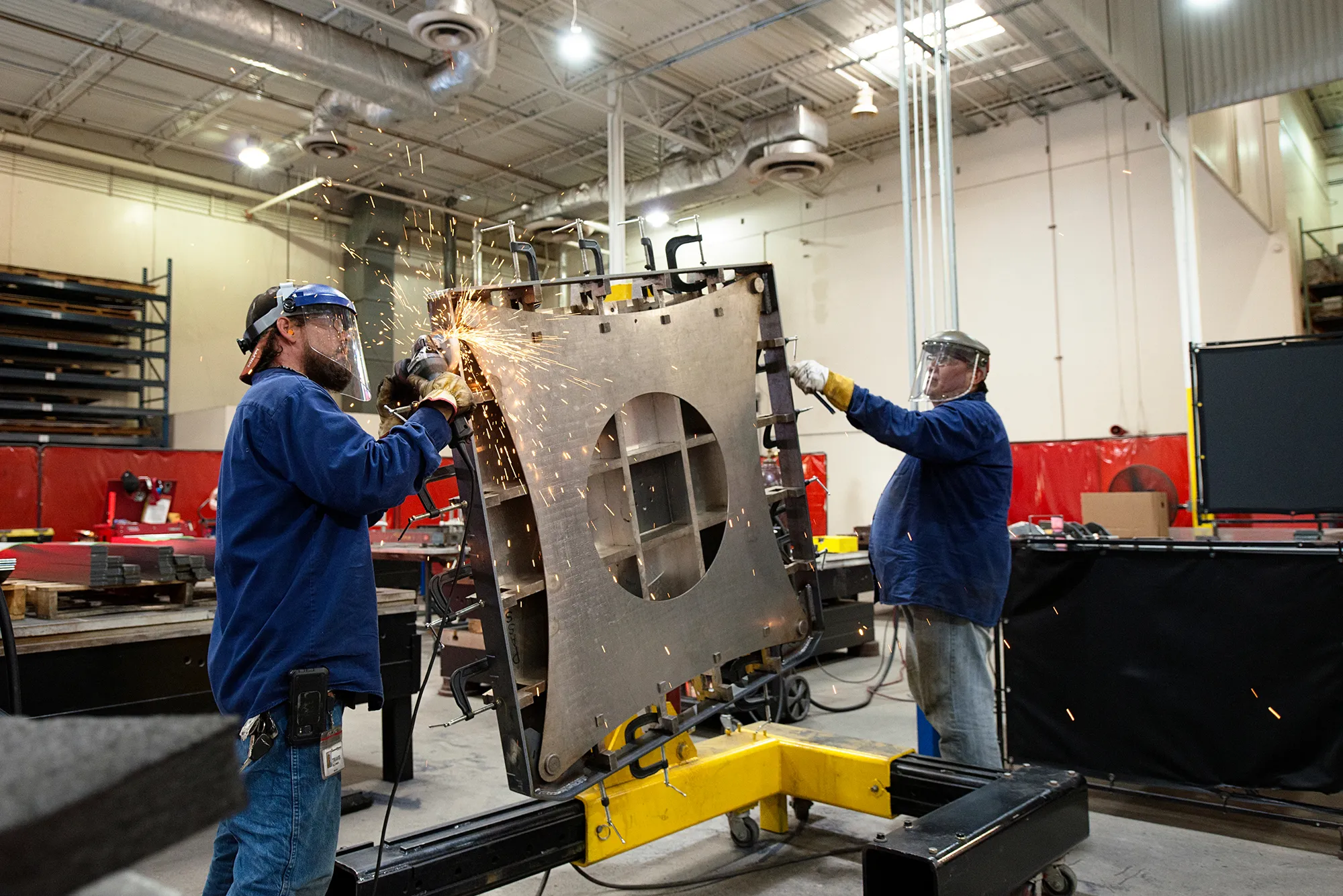 Two workers in protective gear welding and grinding a large metal turret riser inside a factory.