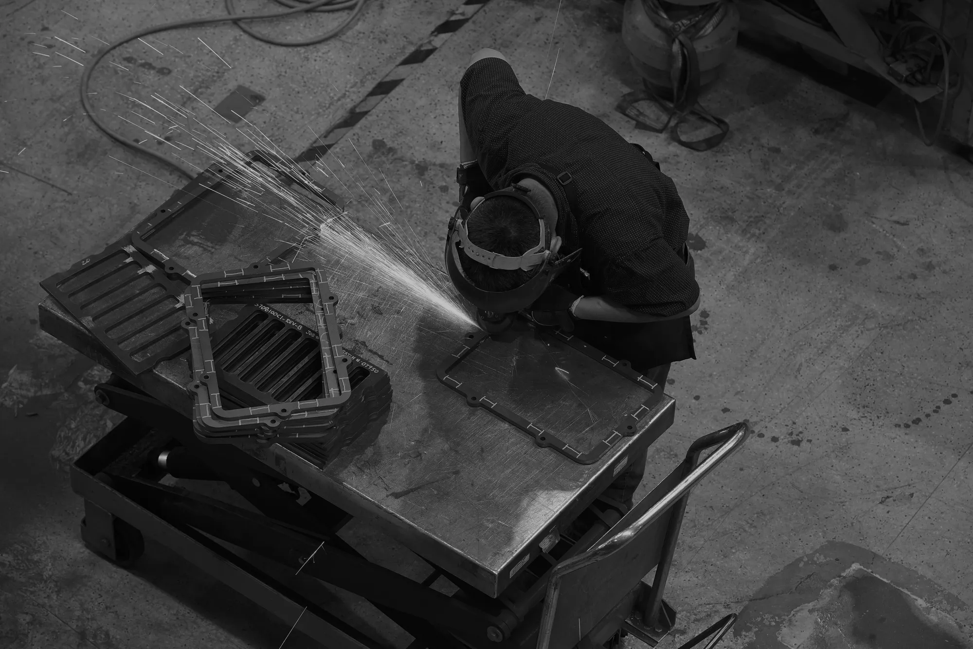 Worker wearing protective gear grinding metal parts on a workbench with sparks flying.