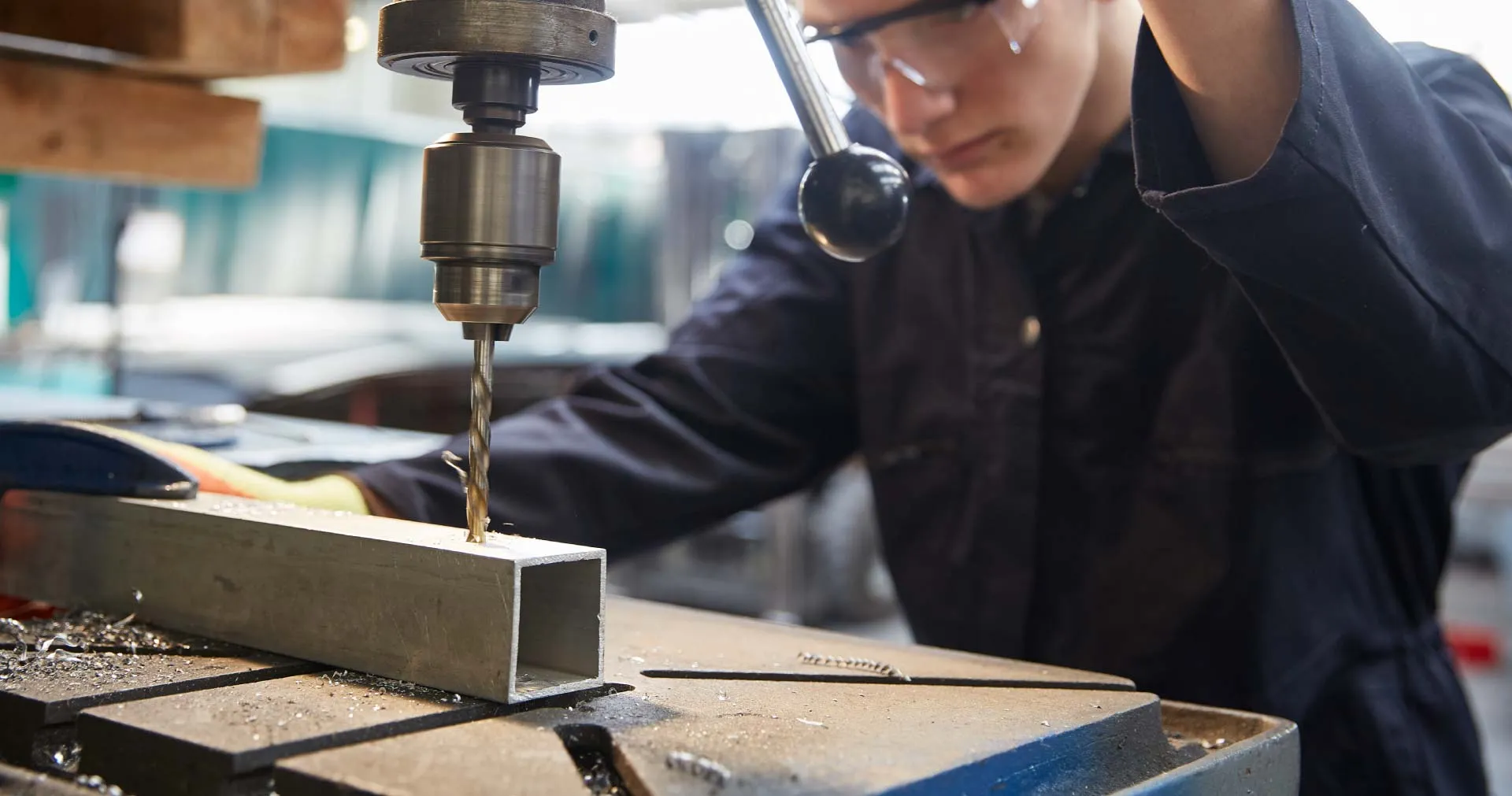 Person wearing safety glasses operating a drill press to drill into a metal square tube.