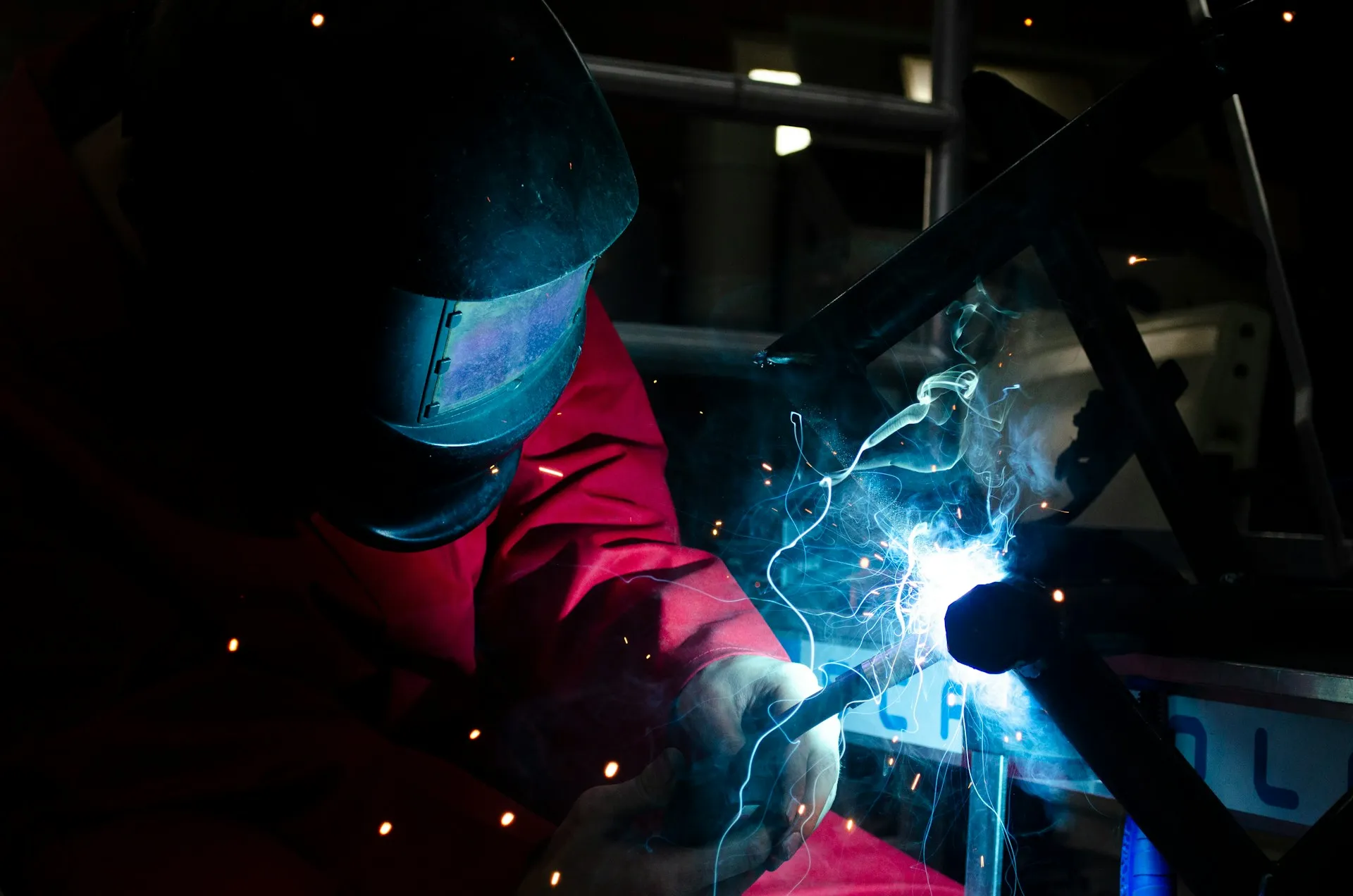 Person welding metal frame with sparks and bright blue light wearing protective gear.
