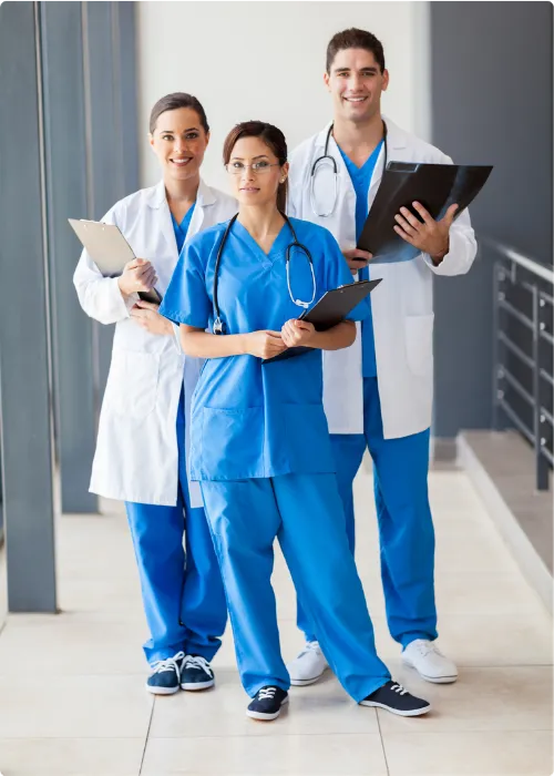 Three medical professionals in blue scrubs and white coats holding clipboards, standing in a hallway.