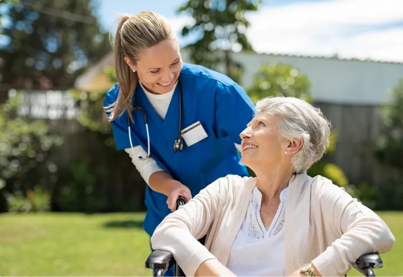 Smiling nurse in blue scrubs pushes elderly woman in wheelchair outdoors in a garden.