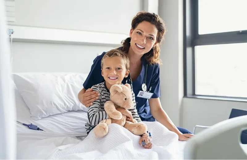 Smiling nurse sitting on a hospital bed with a young boy holding a teddy bear.
