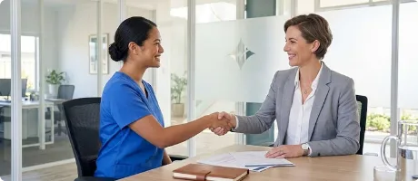 A nurse and a businesswoman smiling and shaking hands across a desk in a modern office.