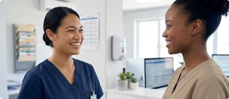 Two healthcare workers smiling and talking in a medical office with computers and files in the background.