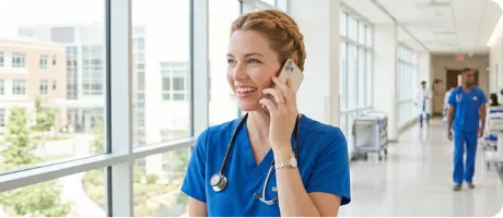 Smiling nurse in blue scrubs talking on a smartphone in a bright hospital corridor.