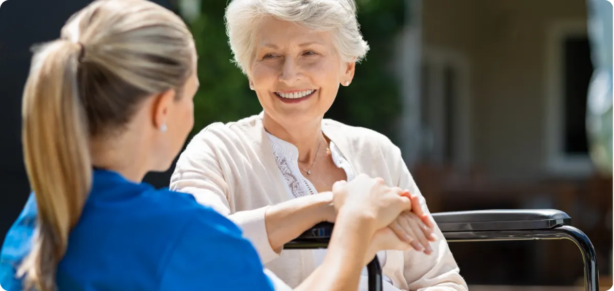 Smiling elderly woman holding hands with a caregiver outdoors near a wheelchair.