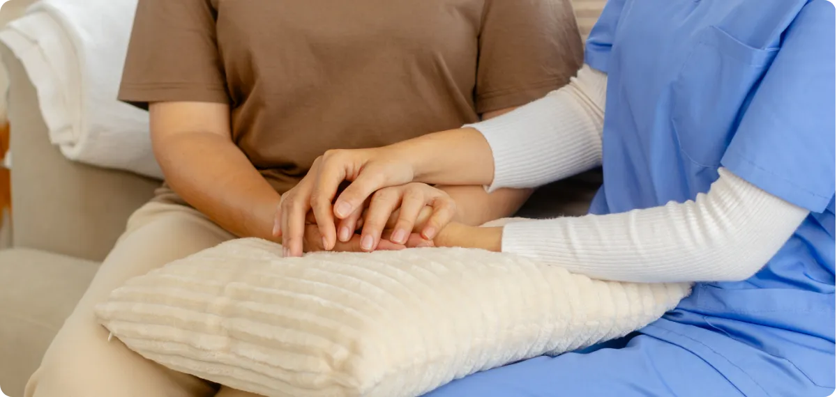 Person in blue scrubs holding hands with a person in brown shirt on a beige pillow.