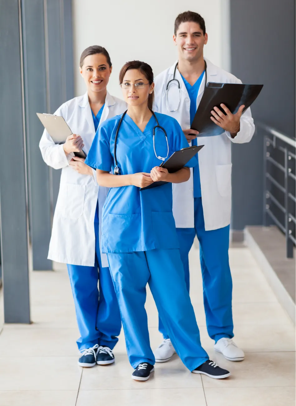 Three smiling medical professionals in scrubs and lab coats holding clipboards and standing in a hallway.