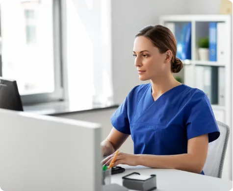 Female healthcare professional in blue scrubs working at a computer in a bright office.