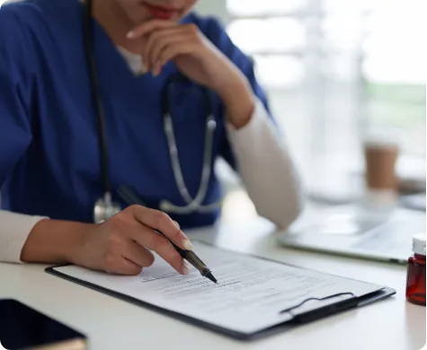 Healthcare professional in blue scrubs writing on a clipboard with a stethoscope around the neck.