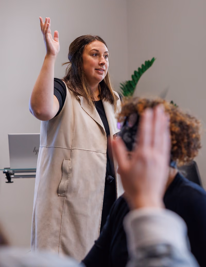 A woman leading a presentation raises her hand in front of a group of attendees