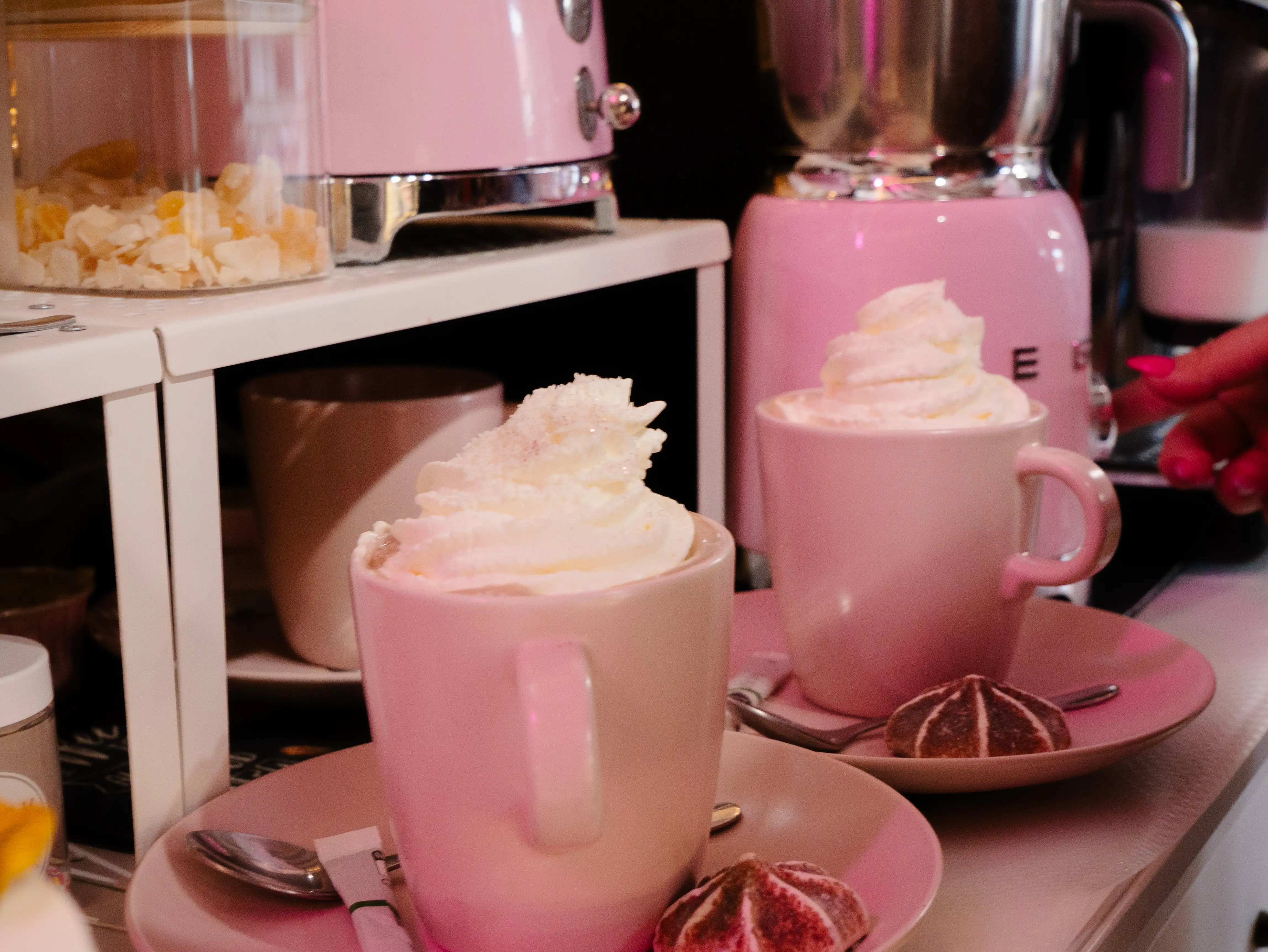 Two pink cups with whipped cream on top, each on a saucer with a small cookie beside it.