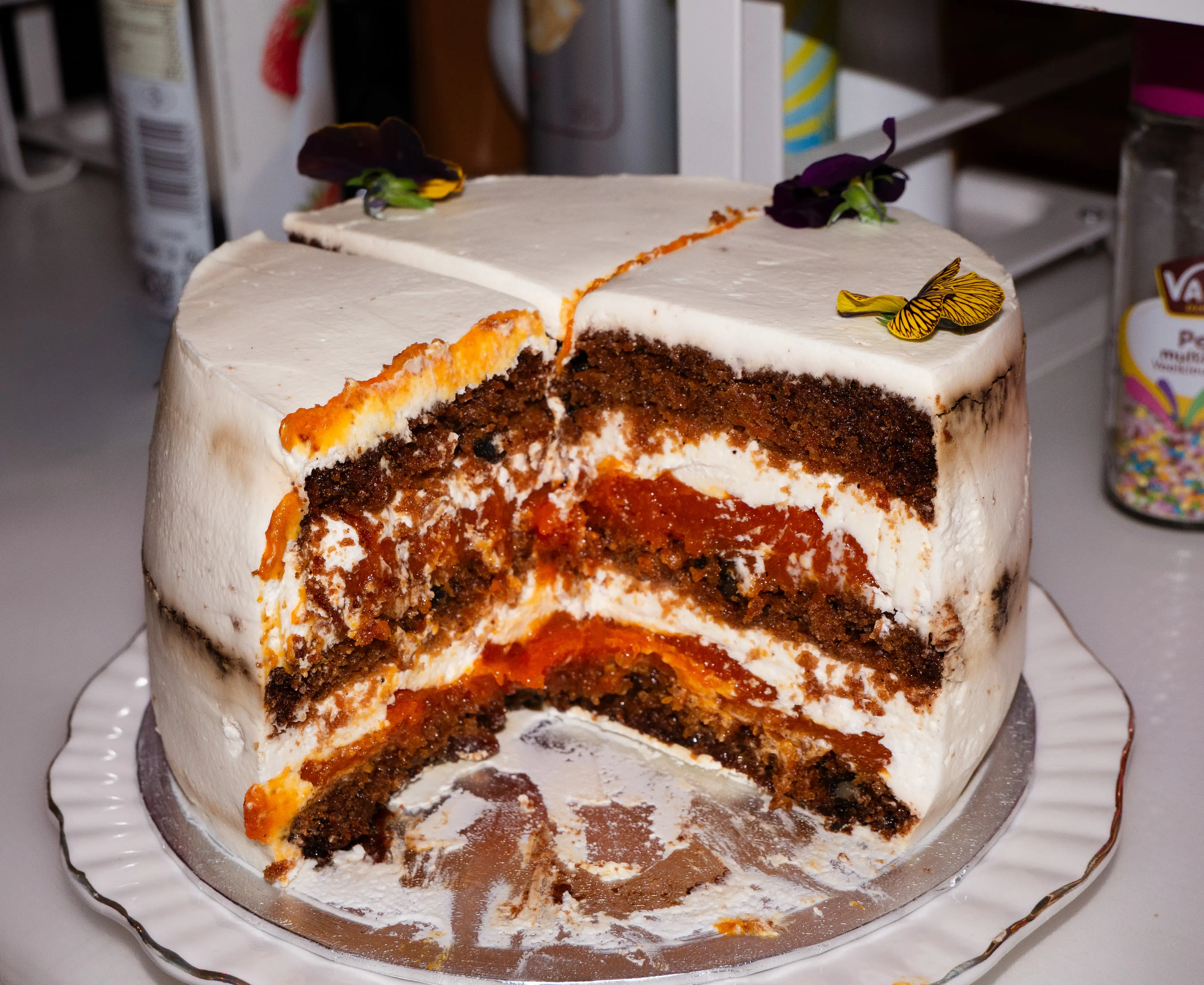 Carrot cake with white frosting and edible purple and yellow flowers on top, partially sliced to show layers.