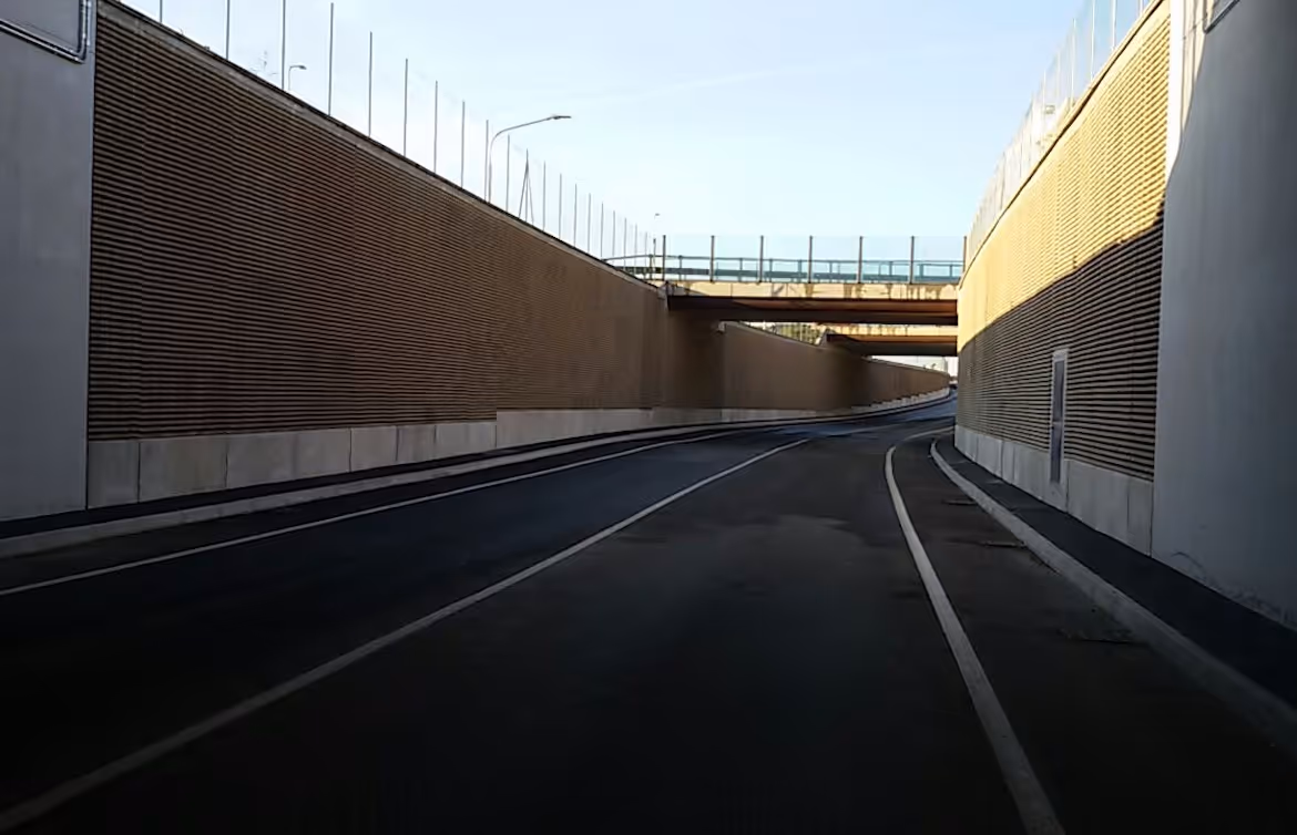 Strada vuota in discesa con muri laterali rivestiti da barriere antirumore e un ponte sopra di essa.