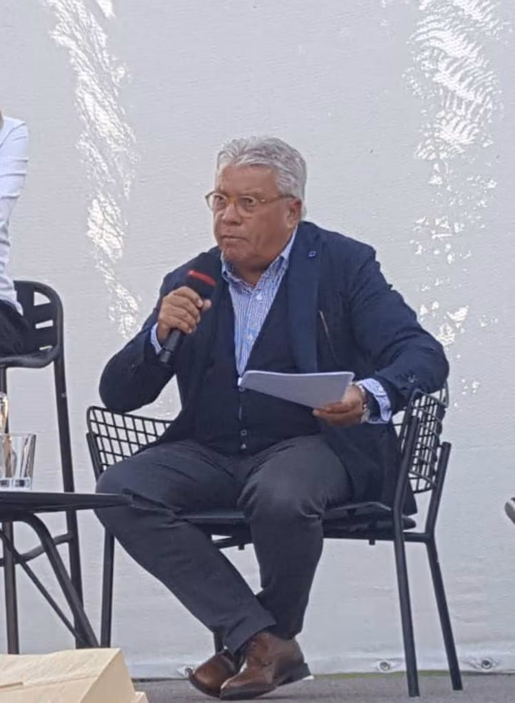 Uomo seduto con microfono e fogli in mano durante un incontro pubblico