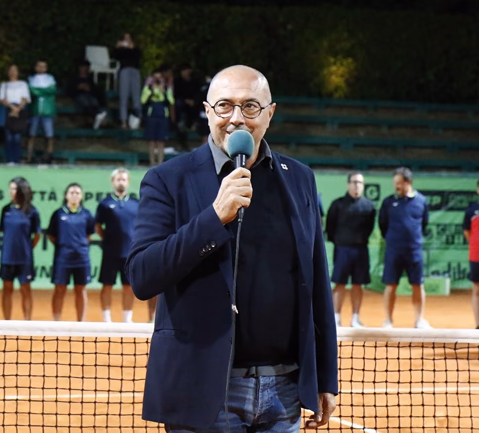 Uomo con il microfono in mano a bordo di un campo da tennis, con i giocatori sullo sfondo