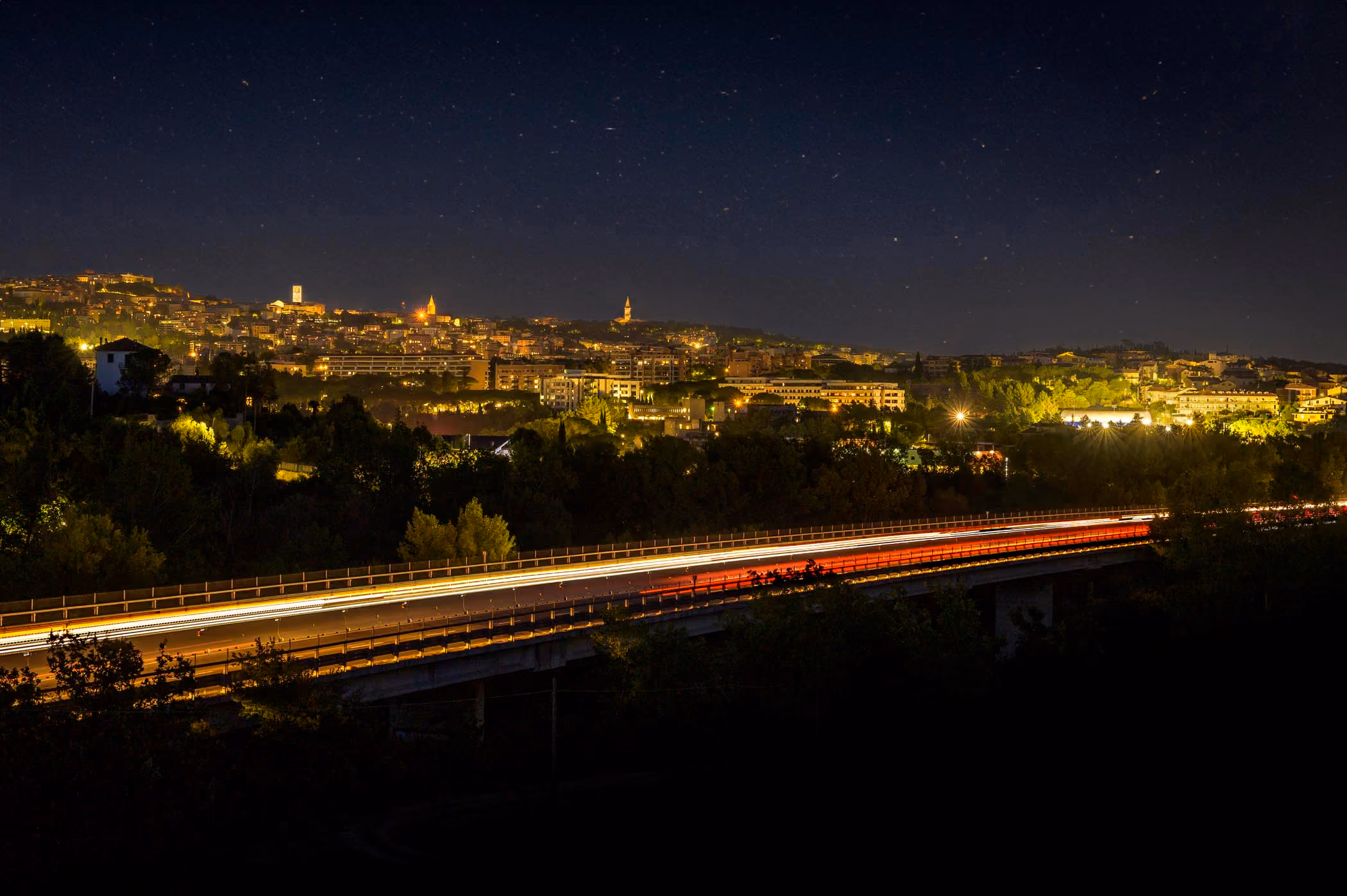 Scia luminosa dei veicoli su un'autostrada di notte, con una città illuminata sullo sfondo