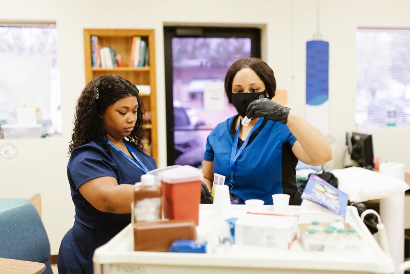 Two Compass Career College Practical Nursing students in blue scrubs preparing medical supplies in a clinical setting.