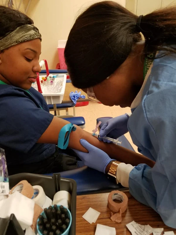 Compass Career College Allied Health student in gloves giving injection to a seated woman's arm using a syringe.