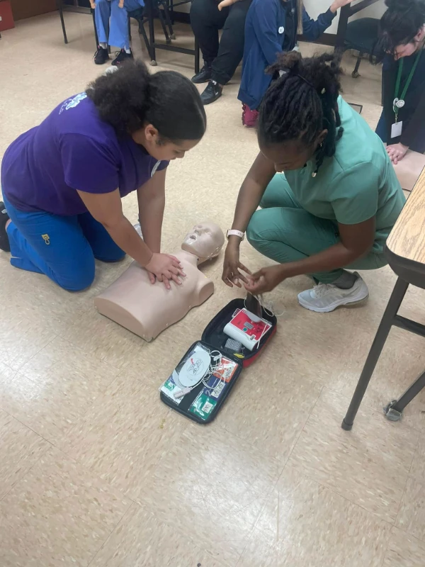 Two Compass Career College Allied Health students practicing CPR on a mannequin with an open AED kit on the floor.